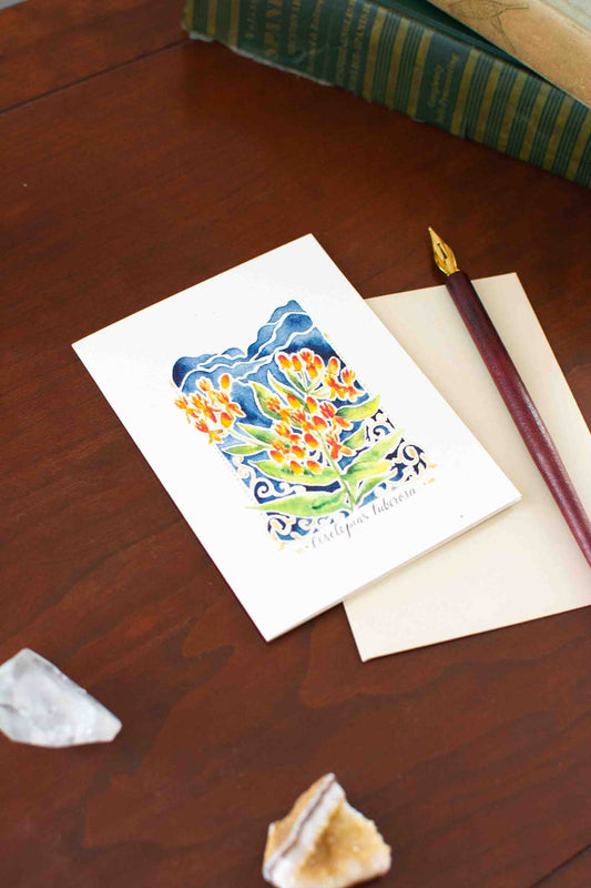 Greeting card with floral Butterfly Weed design on a wooden surface with a pen and envelope.