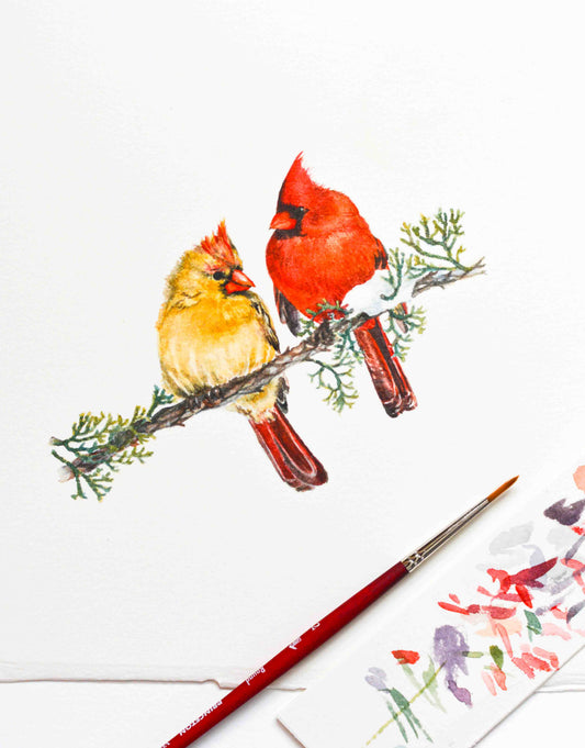 A watercolor painting of two cardinals perched on a branch, shown on white paper with a paintbrush and a small sample of floral paint in the foreground.