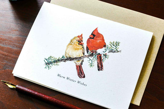 A white greeting card featuring a watercolor illustration of two cardinals on a branch with the text 'Warm Winter Wishes,' shown on a dark wooden desk with a kraft envelope behind it and a fountain pen nearby.