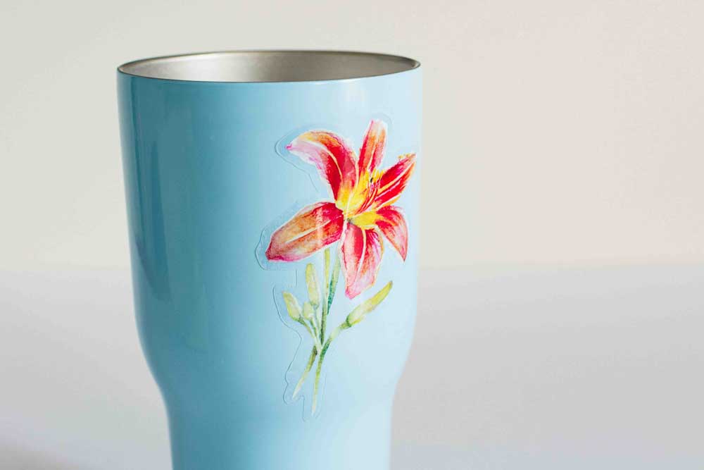 Blue tumbler with a red-orange Daylily sticker on a light gray background