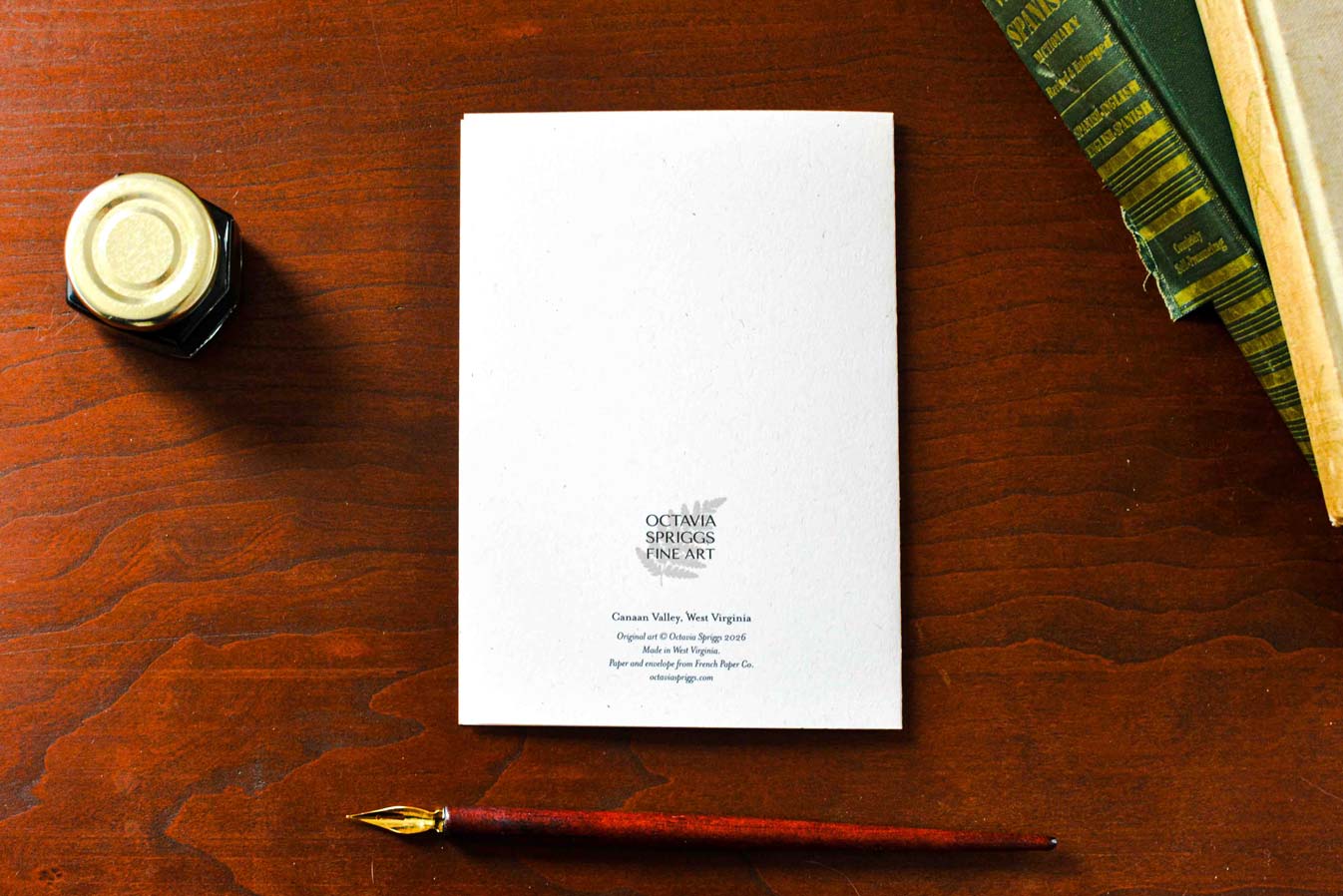 A white card backside with black text reading 'Octavia Spriggs Fine Art' is displayed on a dark wood table, styled with a gold-capped ink bottle, a fountain pen, and books in the corners.