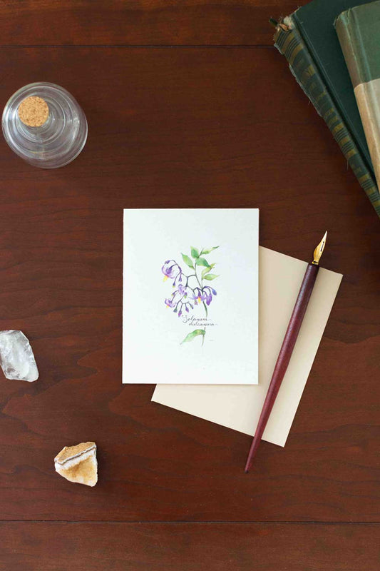 Notebook with floral Nightshade illustration on a wooden surface with a pen and books.