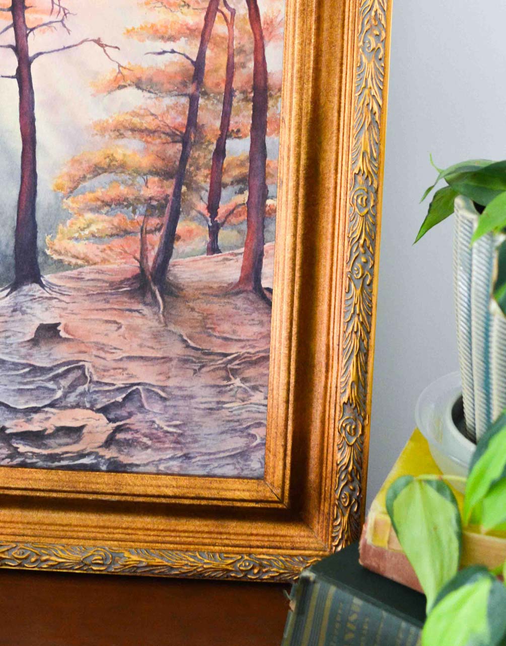 Corner of a painting of an overlook in a gold frame behind a stack of books and plants. 