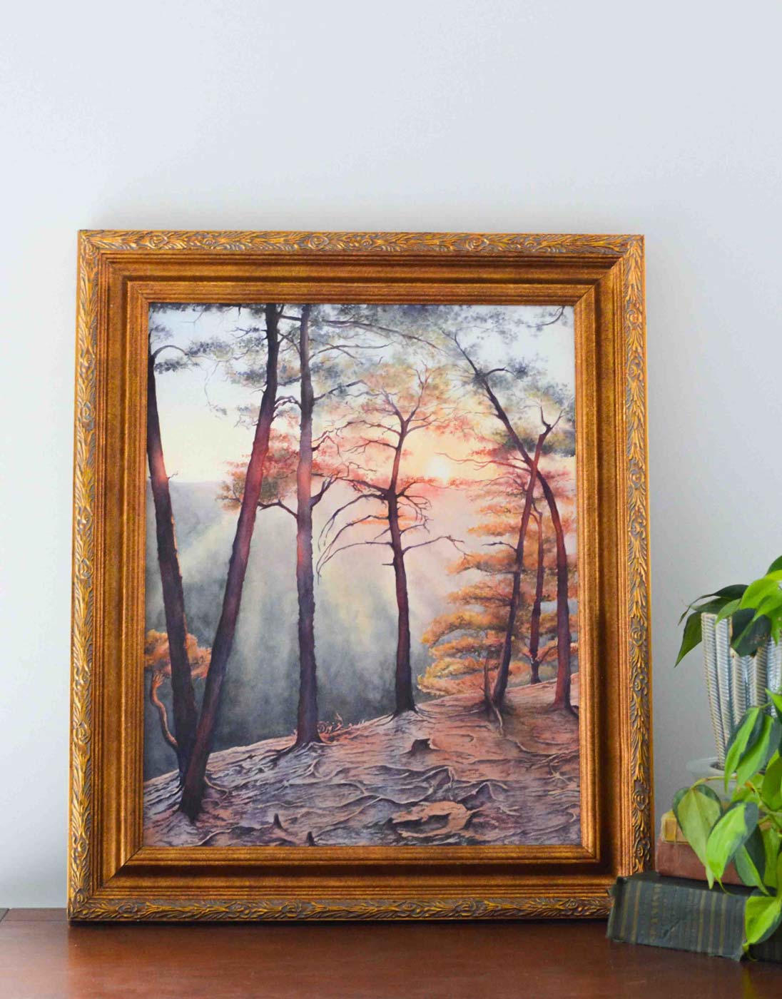 Corner of a painting of Beauty Mountain Overlook in a gold frame behind a stack of books and plants. 