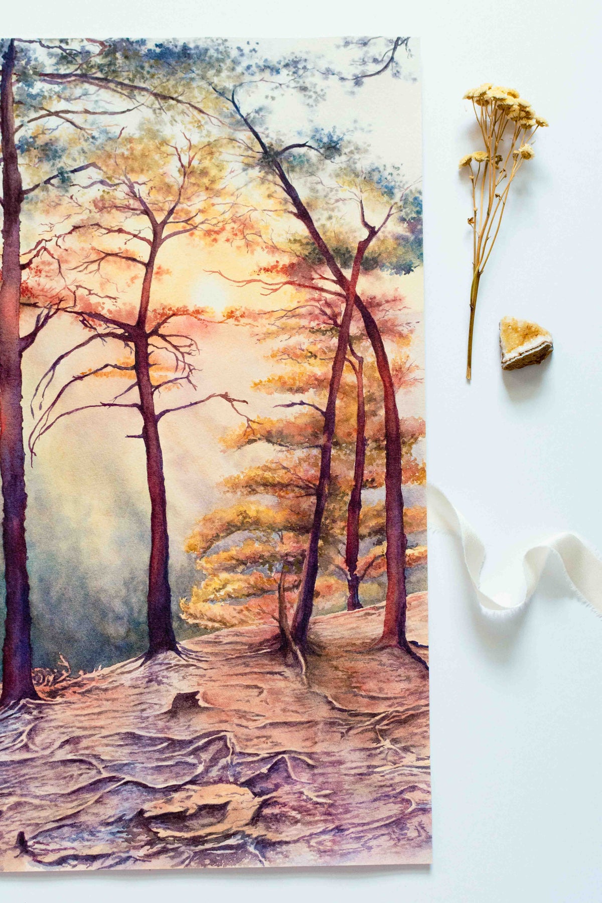 A watercolor painting of a sunset at Beauty Mountain Overlook. Painting shows one side placed next to a citrine gemstone, dried flowers, and ribbon. 