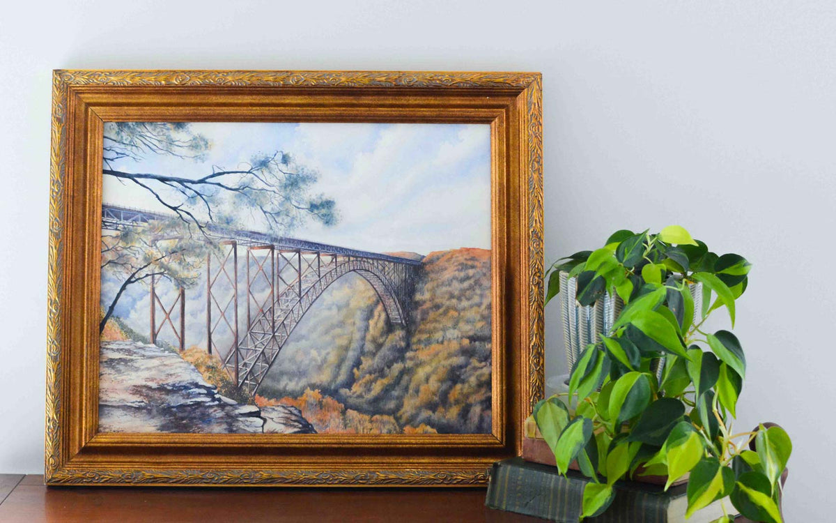 Watercolor painting of the large arched New River Gorge Bridge in a gold frame with a plant nearby on a stack of books. 