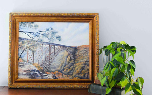 Watercolor painting of the large arched New River Gorge Bridge in a gold frame with a plant nearby on a stack of books. 