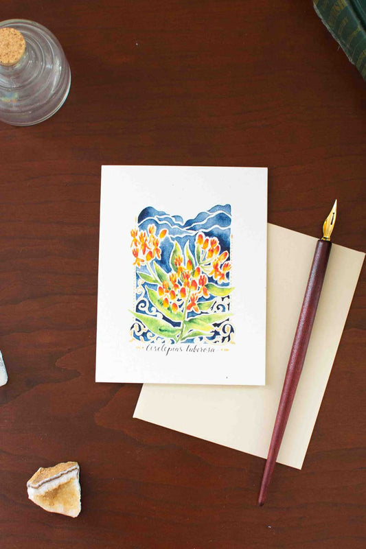 Greeting card with floral Butterfly Weed design on a wooden surface with a pen and envelope.
