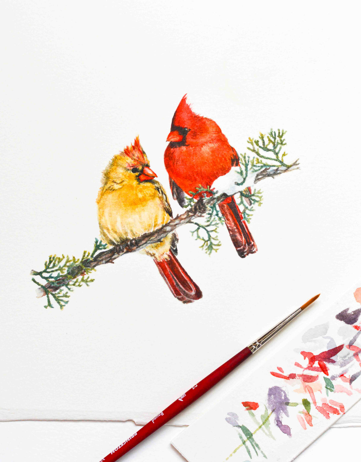 A watercolor painting of two cardinals perched on a branch, shown on white paper with a paintbrush and a small sample of floral paint in the foreground.