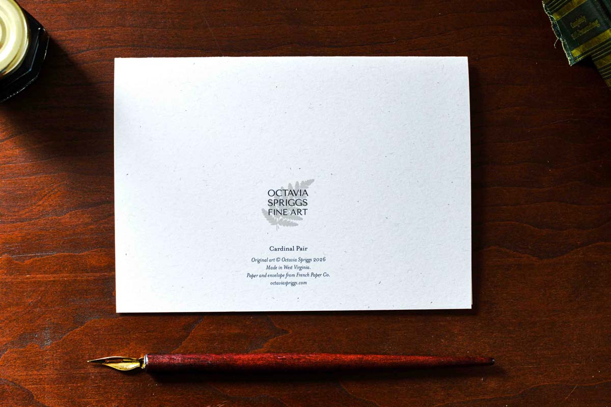 A white art paper card with black text reading 'Octavia Spriggs Fine Art' and 'Cardinal Pair' lies on a dark wooden table, with a red-brown fountain pen placed below it.