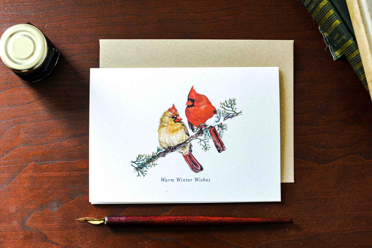 A white greeting card with a watercolor illustration of two cardinals on a branch, printed with the text 'Warm Winter Wishes,' shown with a kraft envelope on a wooden desk. A fountain pen, ink bottle, and books are partially visible around it.