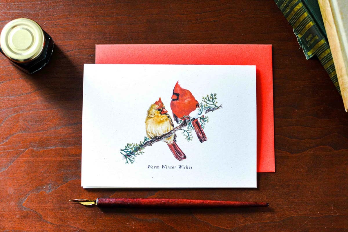 A white greeting card with a watercolor illustration of two cardinals on a branch, printed with the text 'Warm Winter Wishes,' placed on a red envelope over a dark wooden tabletop. A small ink bottle, a paintbrush, and books are partially visible around it.