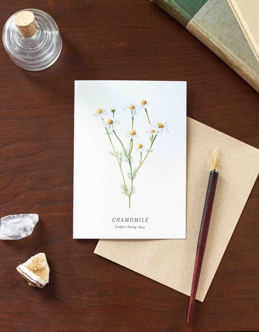 Note card with chamomile illustration on a wooden desk with a pen and crystals.