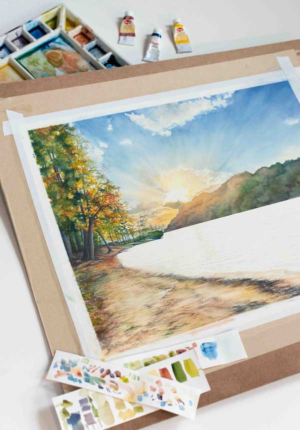 Half-finished painting of a landscape with trees and a river, surrounded by watercolor paints and brushes.