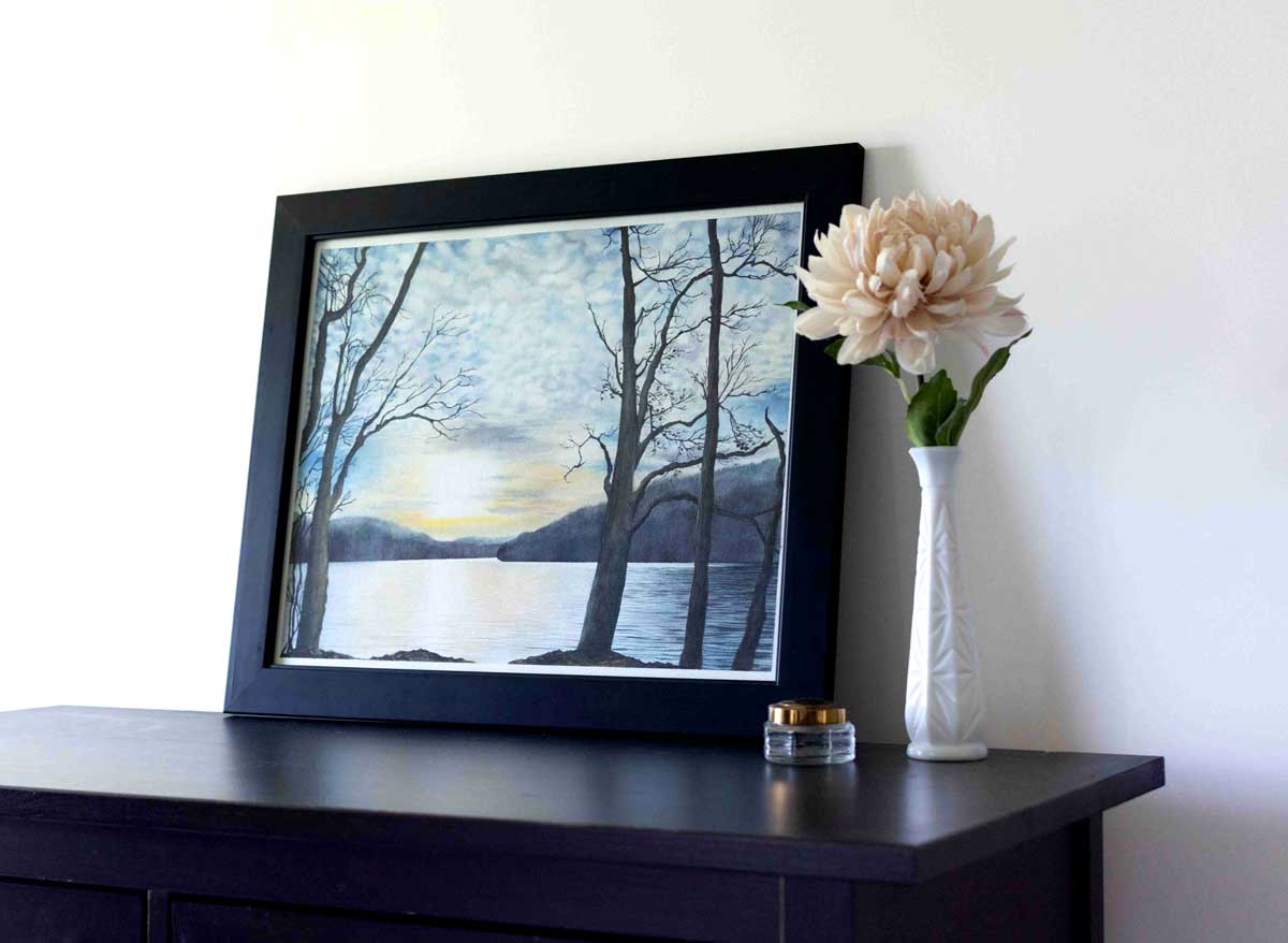 Framed artwork of Cheat Lake with trees and sunset on a table next to a vase with flowers.