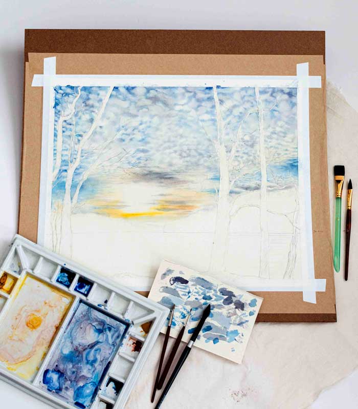 Work in progress of a watercolor painting of a landscape with brushes and paint palette on a light surface.