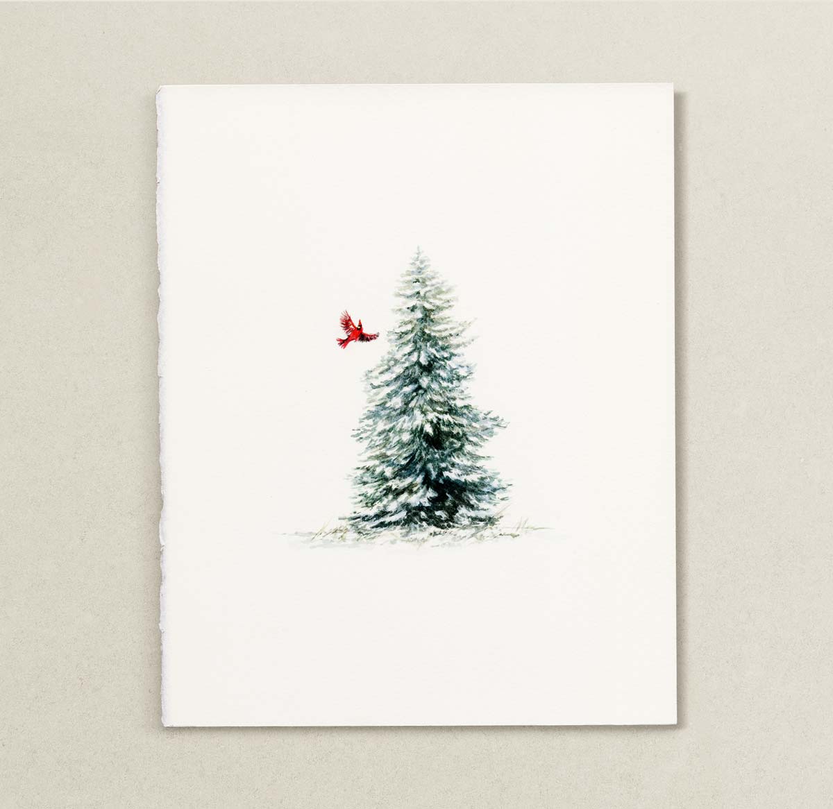 A vertical watercolor painting featuring a snow-dusted evergreen tree with a small red cardinal perched on its side, set against a clean off-white background and displayed on a textured light beige wall.
