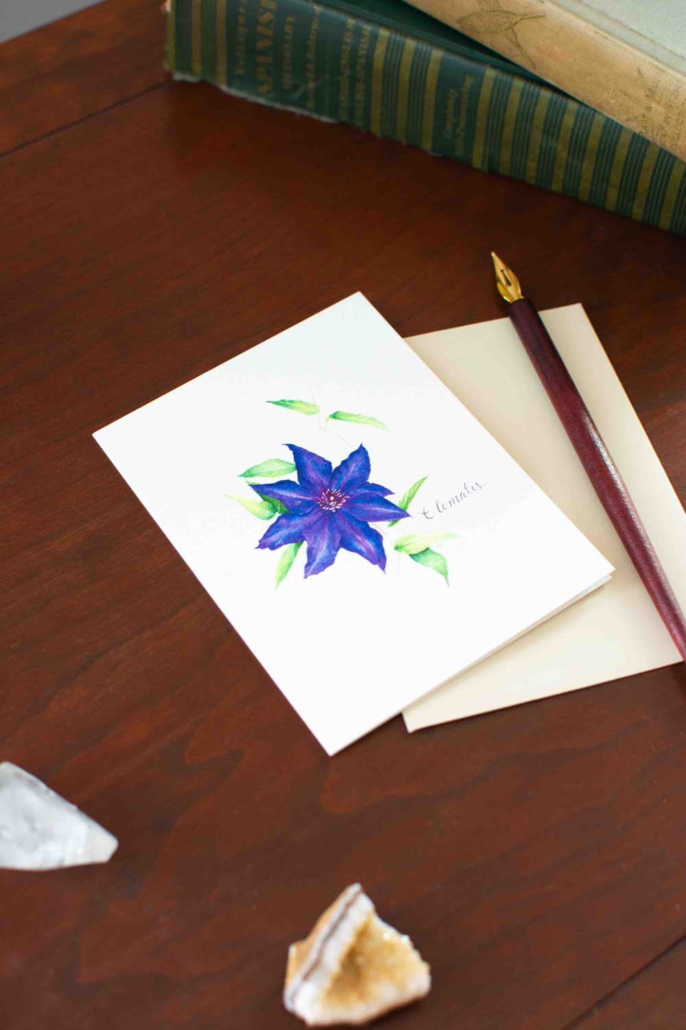Note card with a purple/blue Clematis flower illustration on a wooden surface with a pen and stones.