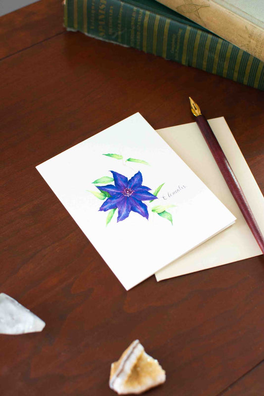 Note card with a purple/blue Clematis flower illustration on a wooden surface with a pen and stones.