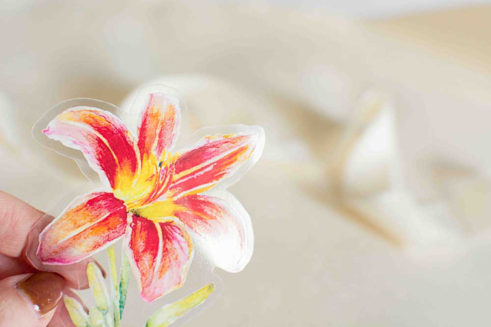 Hand holding a clear vinyl sticker with a red-orange Daylily design on a beige fabric background to show its transparency.