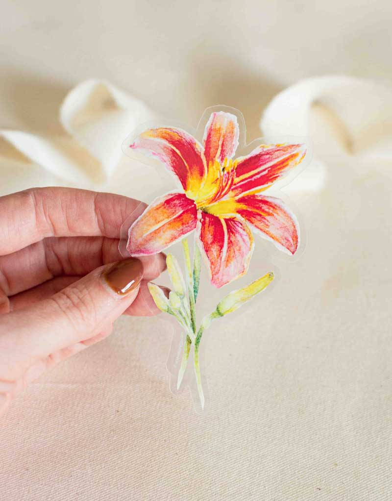 Hand holding a clear vinyl sticker with a red-orange Daylily design on a beige fabric background to show its transparency.