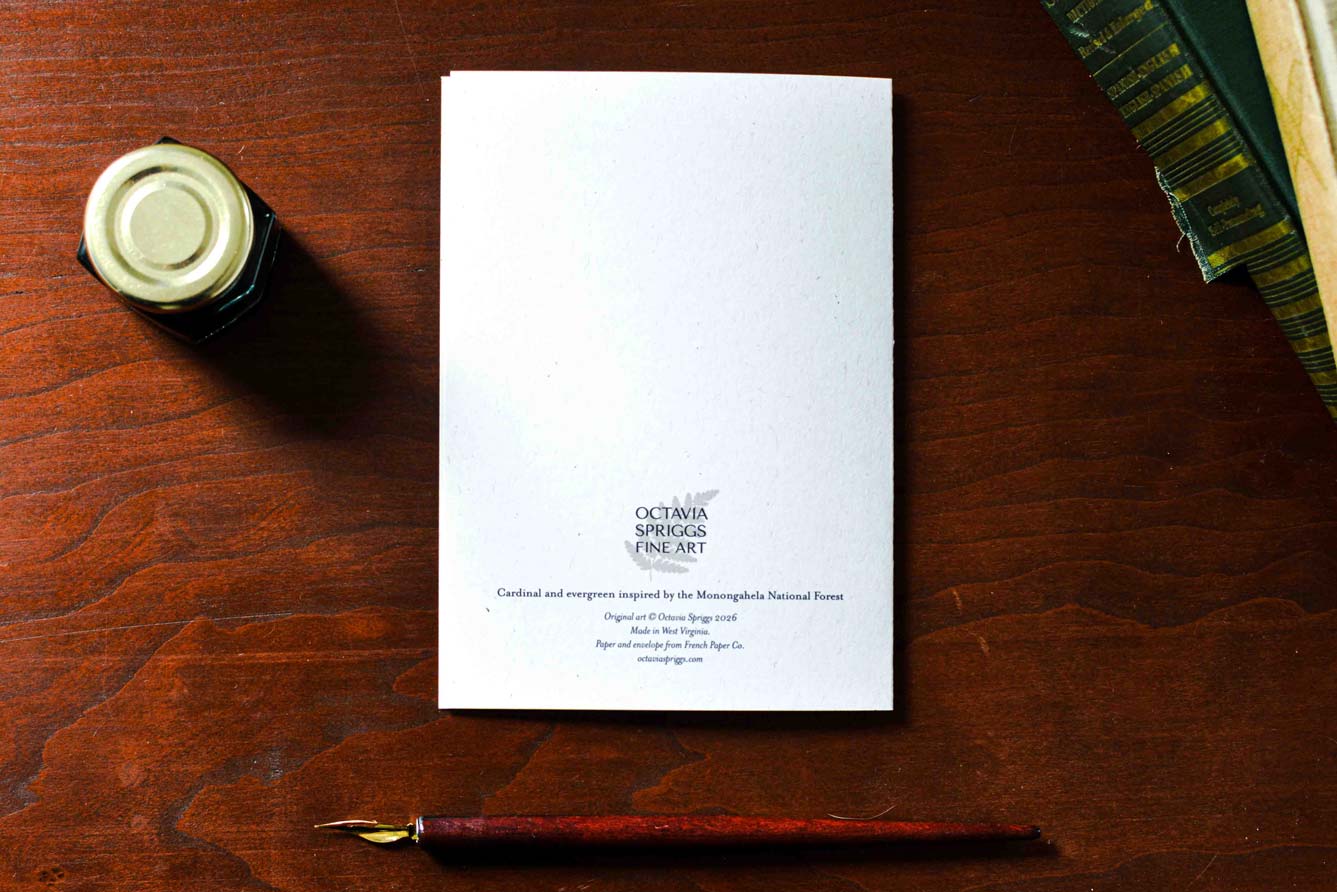 Backside of a white greeting card with the Octavia Spriggs Fine Art logo, styled on a dark wooden desk with a fountain pen, an ink bottle, and books.