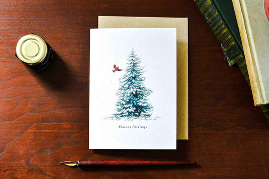 A white Christmas greeting card with a watercolor-style blue evergreen tree and a small red bird, placed on a brown wooden table with a kraft envelope, fountain pen, and books nearby.  Text below the tree reads "Season's Greetings" 