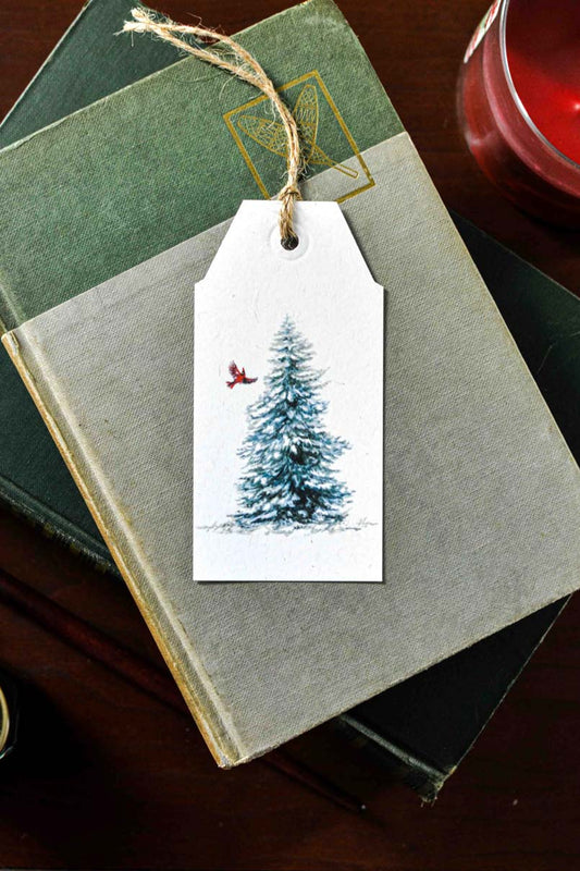 A white gift tag with a printed illustration of a snowy Christmas tree and a small red cardinal, tied with twine and placed on top of stacked books.