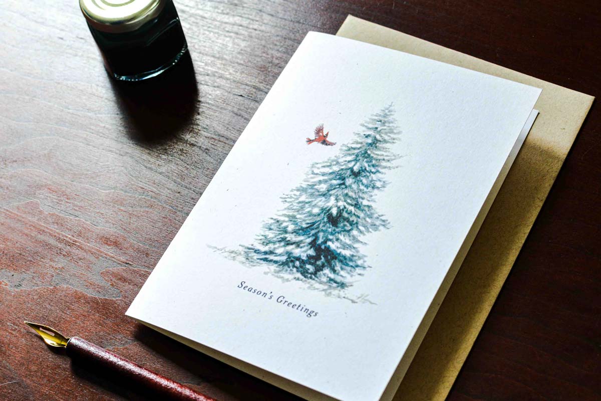 A white Christmas greeting card featuring a watercolor illustration of a snow-covered evergreen tree with a small red bird and the text 'Season's Greetings', placed on a brown kraft envelope over a dark wooden table with a fountain pen and ink bottle nearby.