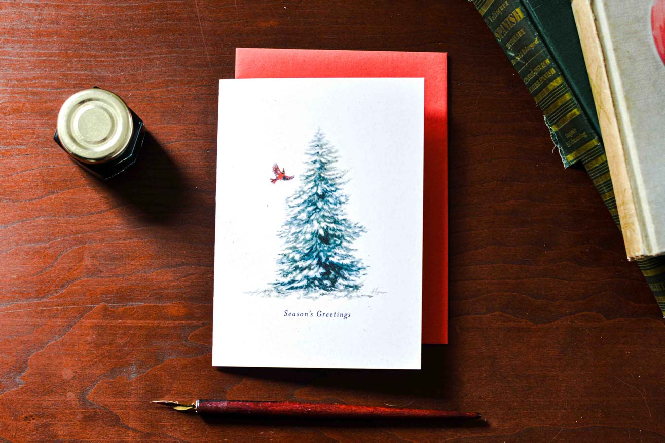 A white greeting card with a blue-green Christmas tree illustration and a small red bird, placed on a red envelope on a wooden table. A small ink bottle, fountain pen, and stacked books are nearby.