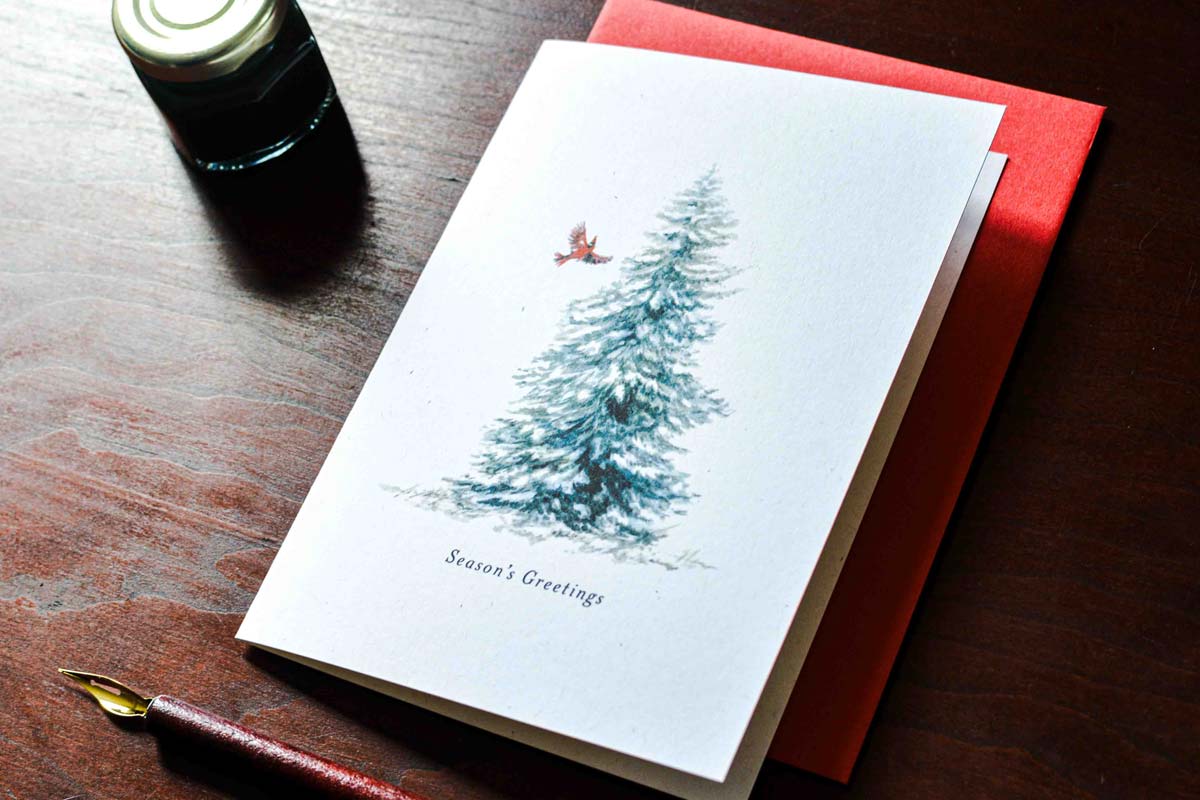 A white greeting card with a watercolor illustration of a snow-covered evergreen tree and a small red bird, reading 'Season's Greetings,' placed on a dark wooden table with a red envelope behind it.