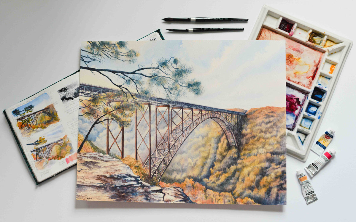Watercolor painting of the large arched New River Gorge Bridge laying on top of a sketchbook and a watercolor palette with brushes and paints nearby. 