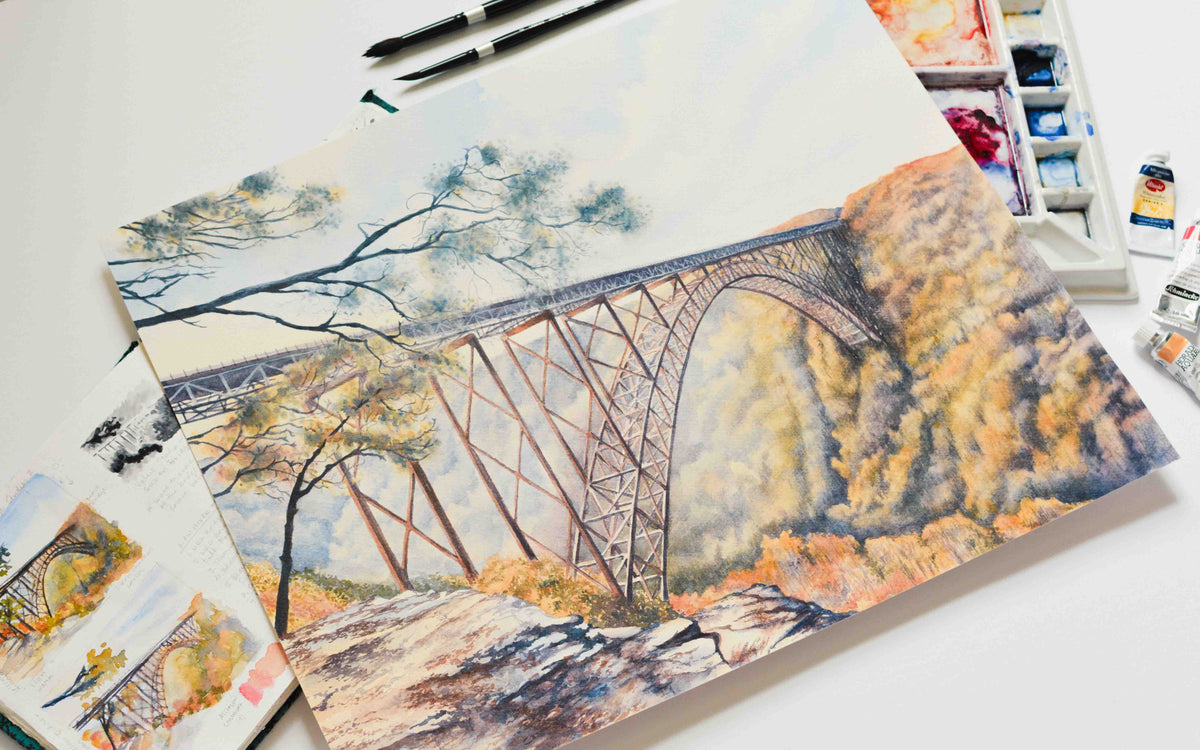 Watercolor painting of the large arched New River Gorge Bridge laying on top of a sketchbook and a watercolor palette with brushes and paints nearby. 