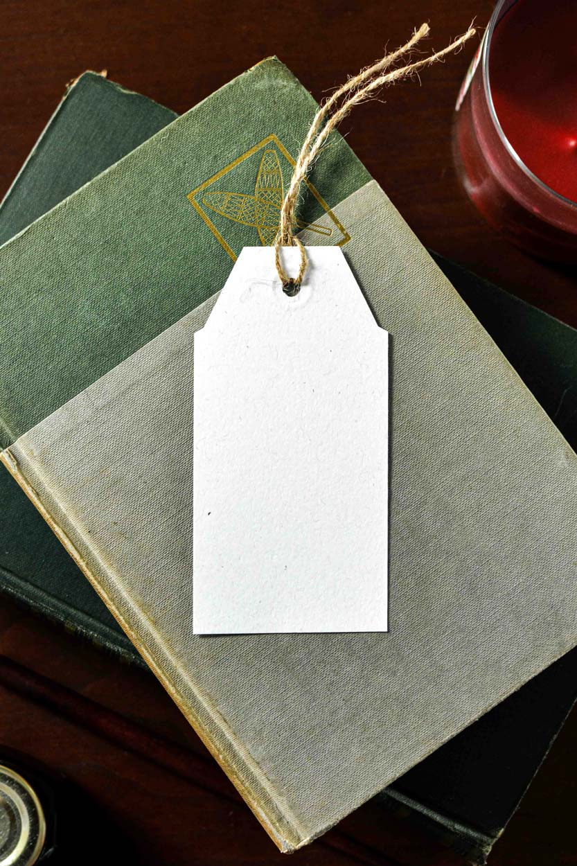 A blank white paper gift tag with a rustic twine loop, laid on top of a gray-green book with another green book underneath on a dark wooden surface.
