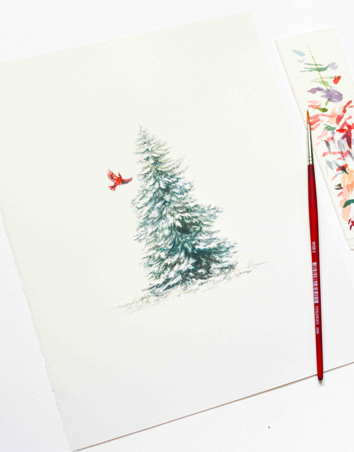 A watercolor painting of a snow-covered evergreen tree with a small red cardinal on a white sheet of paper, styled with a red paintbrush and a floral art card on a white surface.
