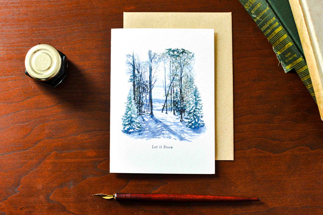 A white greeting card with a blue watercolor winter forest illustration and the words 'Let it Snow' lies on a brown wooden table, partially covering a kraft envelope. A small ink bottle, fountain pen nib, and stacked books are arranged around it as styled props.