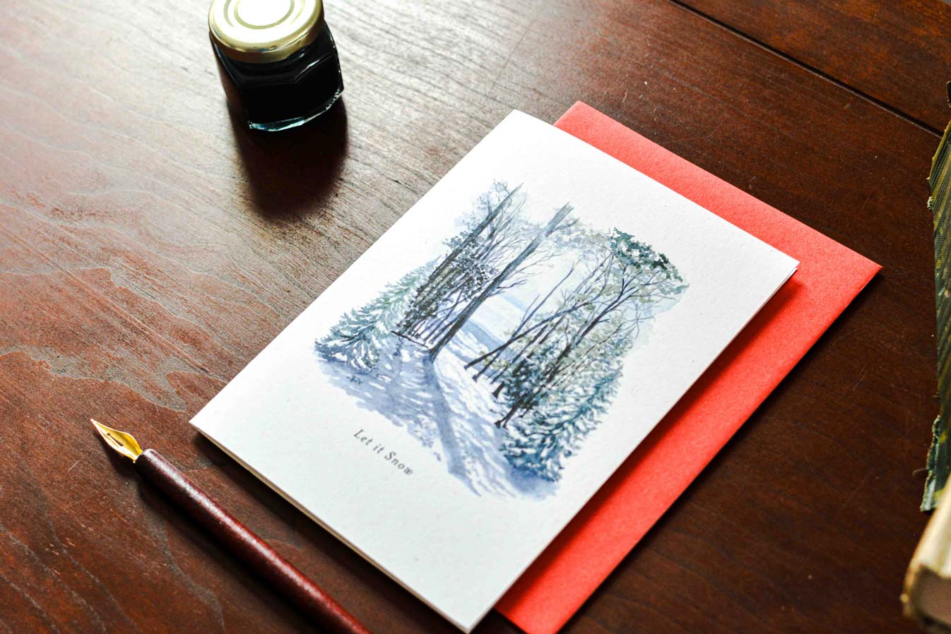 A card featuring a blue-gray winter forest scene with a snowy path, placed on a red envelope over a dark wooden table. A fountain pen and a small ink bottle are also visible in the scene.