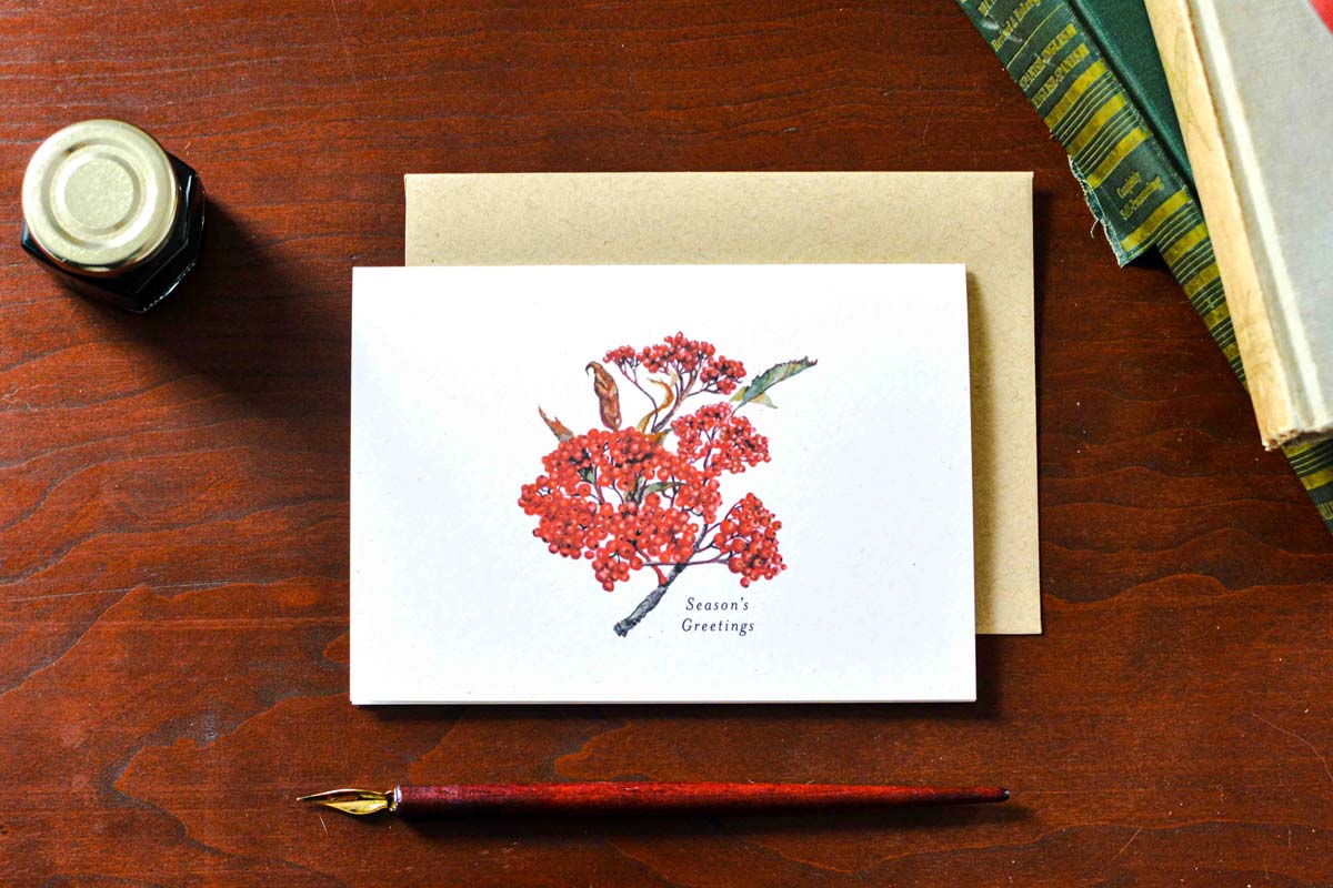 A white greeting card with a watercolor illustration of red berries and small birds, placed on a kraft envelope on a wooden table. A fountain pen, ink bottle, and books are visible around it. Text below the berries reads "Season's Greetings" 
