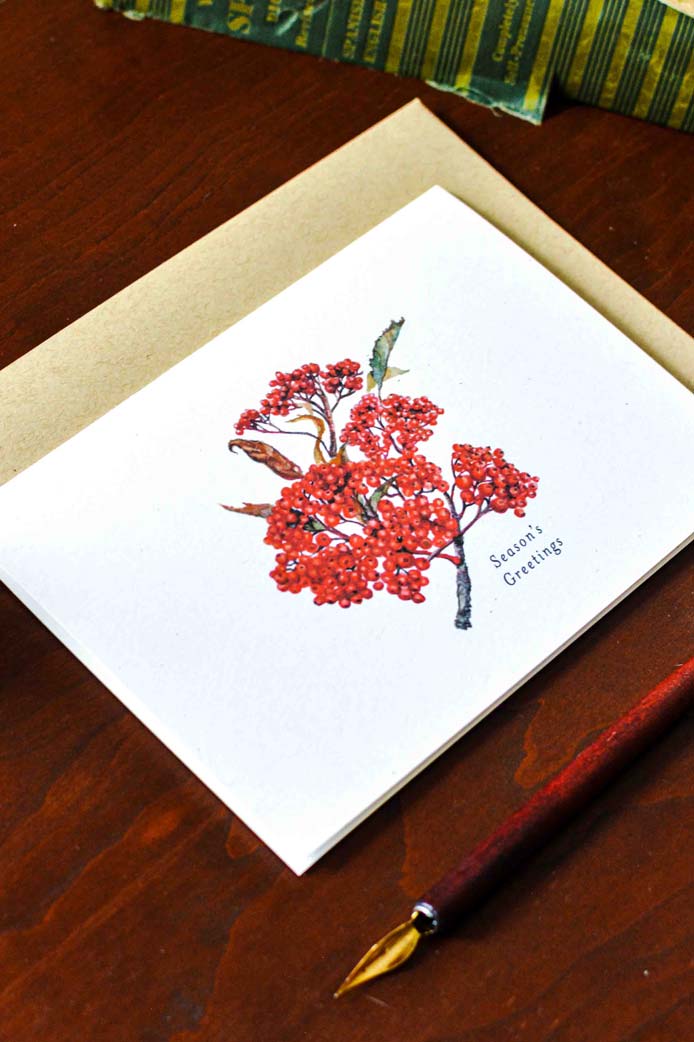 A white greeting card with a red berry illustration and the words 'Season's Greetings' lies on a brown kraft envelope over a dark wooden table, with a gold pen nearby.