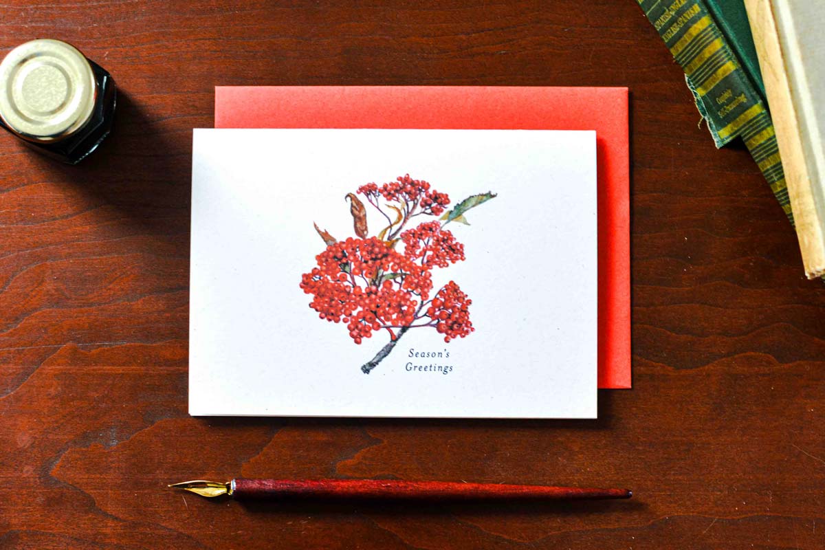 A white greeting card with a red berry branch illustration and the words 'Season's Greetings' is placed on a red envelope over a wooden desk, with a fountain pen, ink bottle, and books nearby.