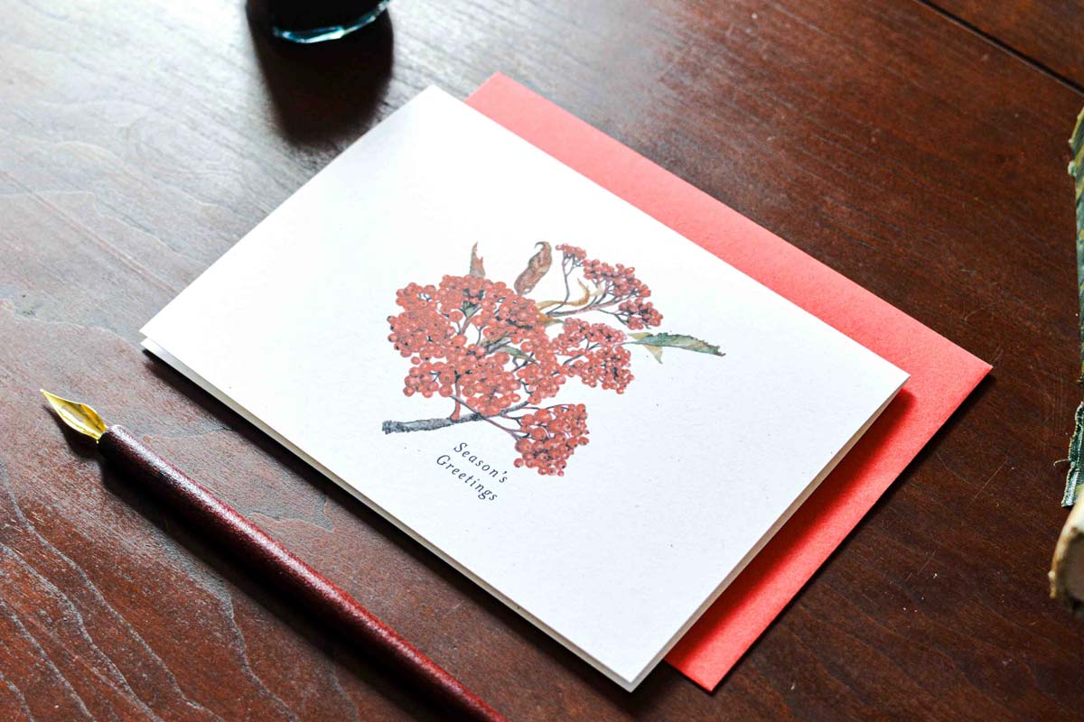 A white greeting card with a delicate watercolor illustration of red berries and birds, placed on top of red envelope on a dark wooden table. A gold-tipped pen lies nearby.