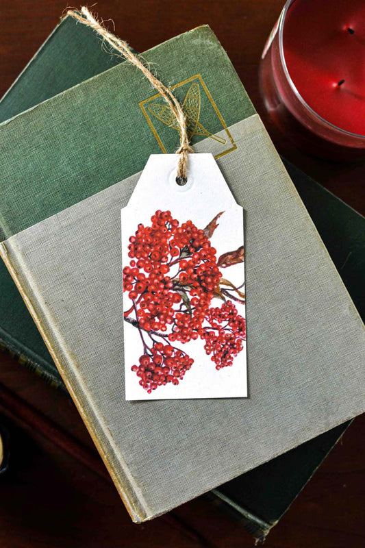 A white gift tag printed with a red berry illustration and tied with twine, resting on stacked vintage books with a red candle partially visible in the background.