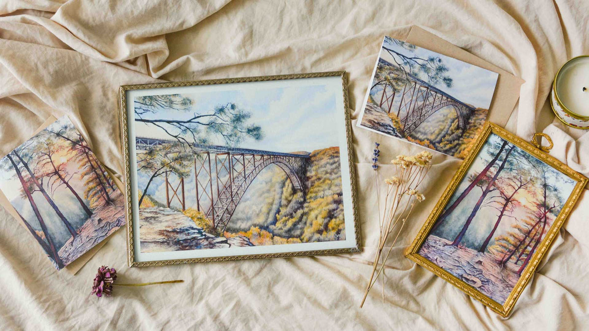 A styled flat-lay of three framed and unframed landscape art prints depicting a the New River Gorge bridge and trees, arranged on beige linen fabric with dried flowers and a candle.