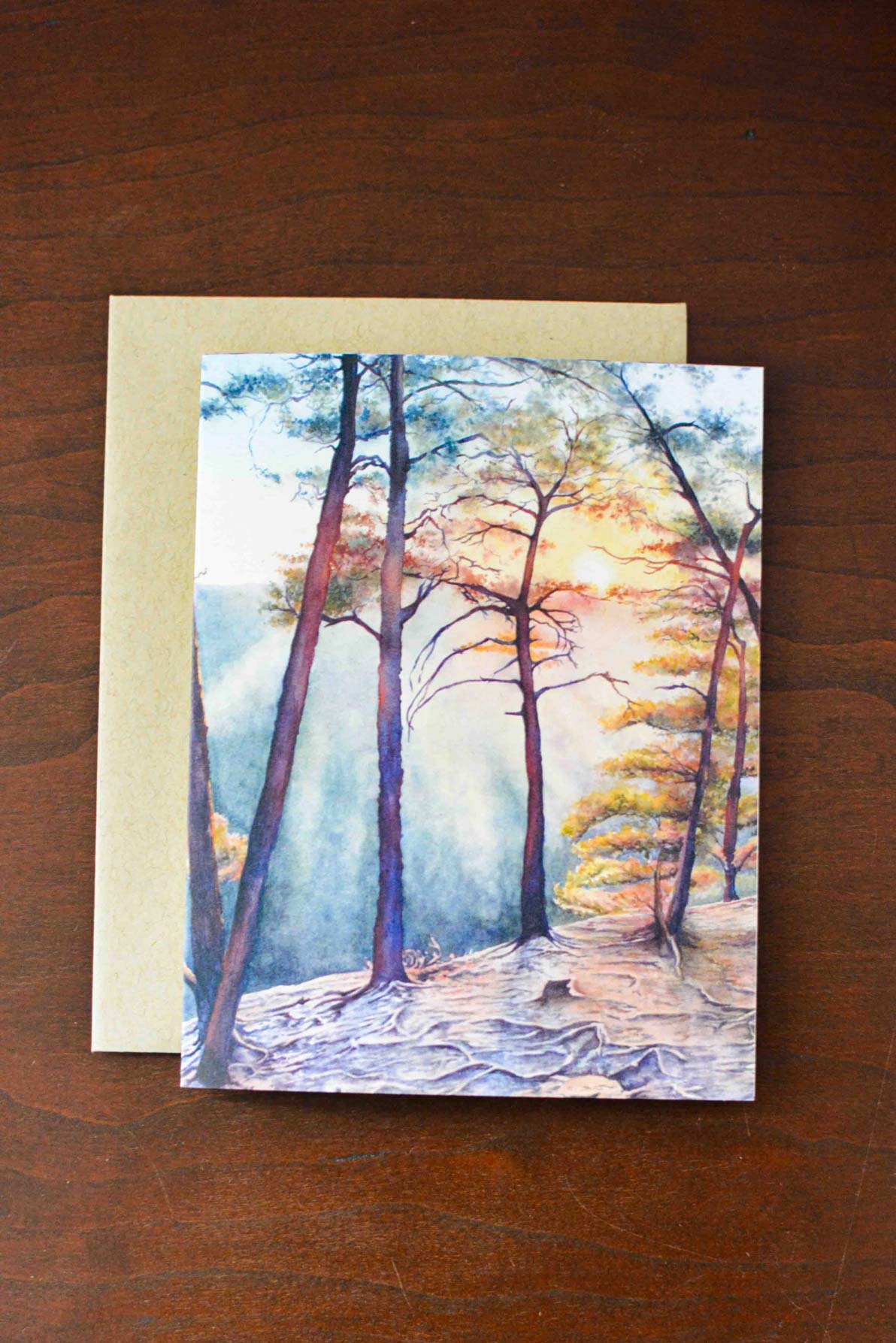 Notecard with a watercolor painting of a sunset at Beauty Mountain with trees. Notecard is laying on a kraft envelope on a wooden desk. 
