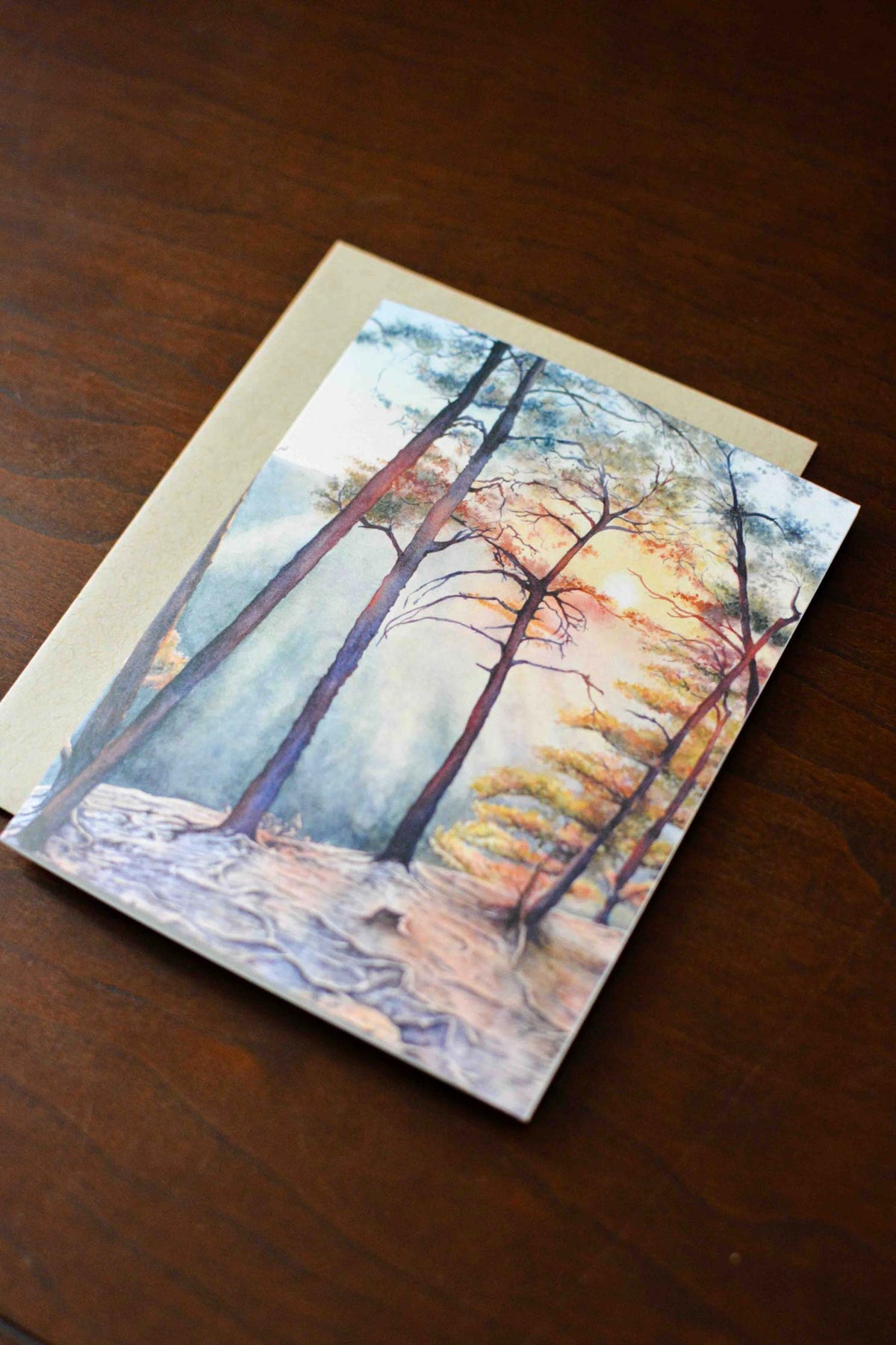 Notecard with a watercolor painting of a sunset at Beauty Mountain with trees. Notecard is laying on a kraft envelope on a wooden desk. 