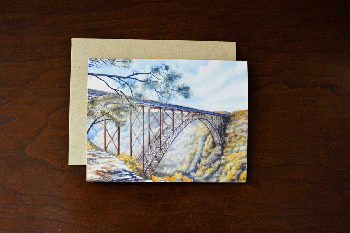 A watercolor note card of the large arched New River Gorge Bridge. The notecard is laying on a desk with a kraft envelope. 