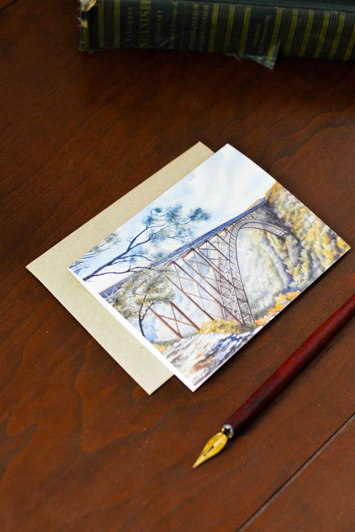 A watercolor note card of the large arched New River Gorge Bridge. The notecard is laying on a desk with a kraft envelope and a calligraphy pen. 