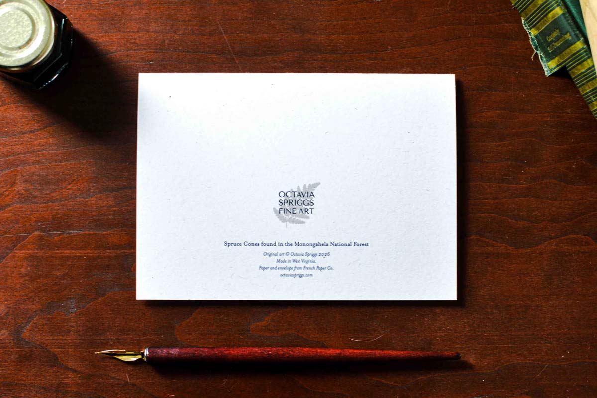 A white art print card featuring the text 'OCTAVIA SPRIGGS FINE ART' and smaller descriptive text, placed on a dark wooden table with a fountain pen and ink bottle nearby.