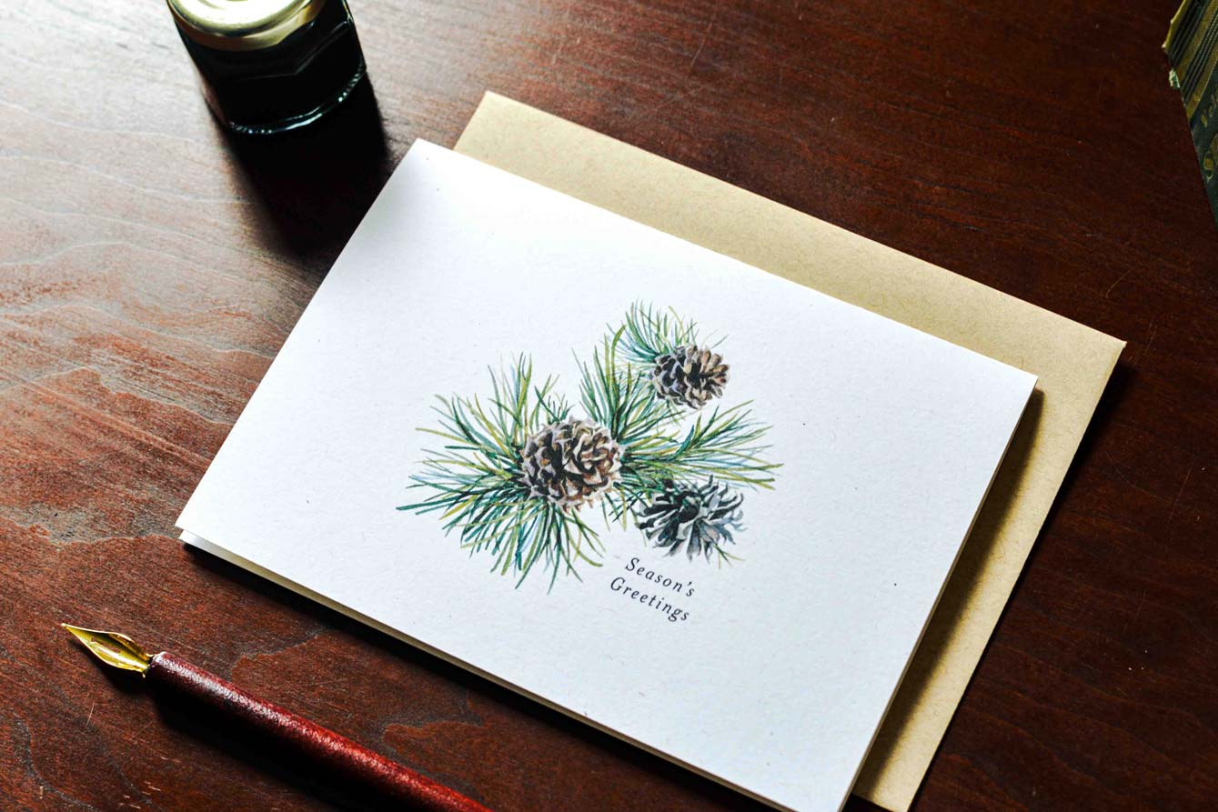 A white greeting card with a watercolor illustration of pine needles and pinecones, with the text 'Season's Greetings,' placed on a kraft envelope on a dark wooden table. A fountain pen and a small bottle are partially visible around it.