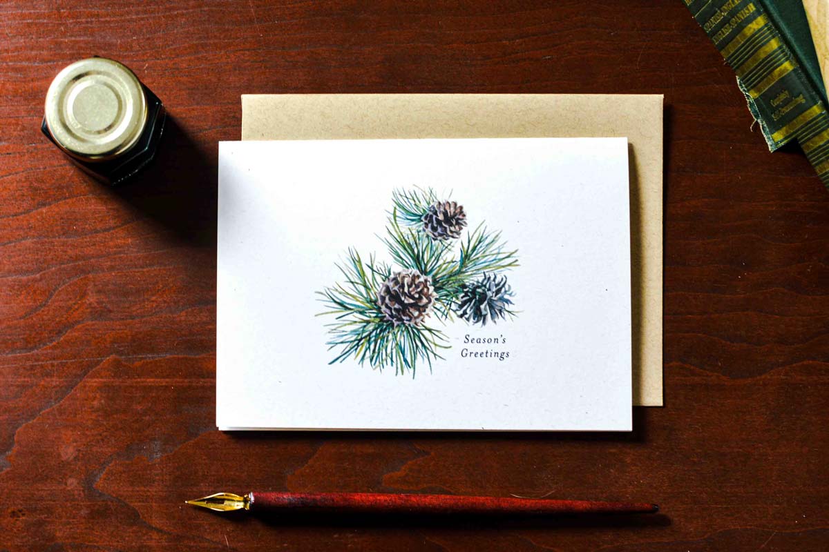 A white greeting card with a watercolor pine branch and pinecone illustration, placed on top of a kraft envelope on a dark wooden table. A fountain pen, a small bottle, and part of a book are visible as styled props.Text below the cones reads "Season's Greetings" 